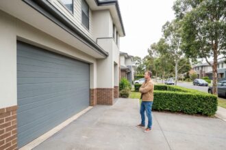 Un homme devant son garage à double porte sectionnelle dans un quartier résidentiel, réfléchissant au choix d'une porte de garage pratique au quotidien