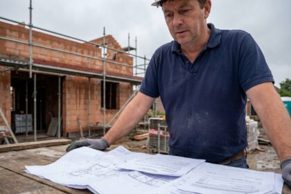 Un homme compare des devis de construction sur un chantier en Belgique avec des plans étalés sur une table en bois